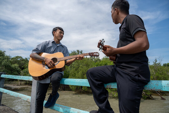Two friends leaning on bridge railing playing acoustic guitar and ukulele. Two musicians enjoying playing music together on a coastal bridge.