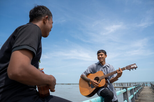 Two friends leaning on bridge railing playing acoustic guitar and ukulele. Two musicians enjoying playing music together on a coastal bridge.