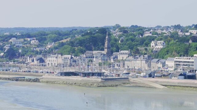 Wide shot of the coastal town of Binic in Brittany, France, showing the marina filled with sailboats, historic stone buildings, and lush green hills in the background.