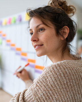 Woman smiles while marking a colorful agenda board. She wears a soft, knitted sweater and has her hair in a bun. Her expression suggests focus and satisfaction. This image captures a creative