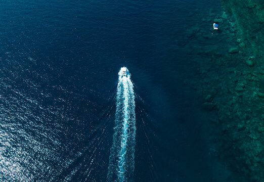 Aerial drone view of a speedboat creating a white wake across deep blue sea near a rocky reef in clear tropical water.