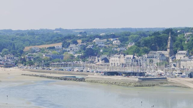 Wide aerial panning shot of Binic, a coastal town , showing the marina with numerous sailboats, the sandy beach at low tide, the town's historic buildings in Brittany, France.