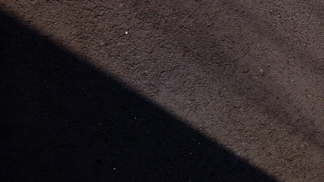 Dark rough asphalt road surface texture split by diagonal sunlight shadow.
