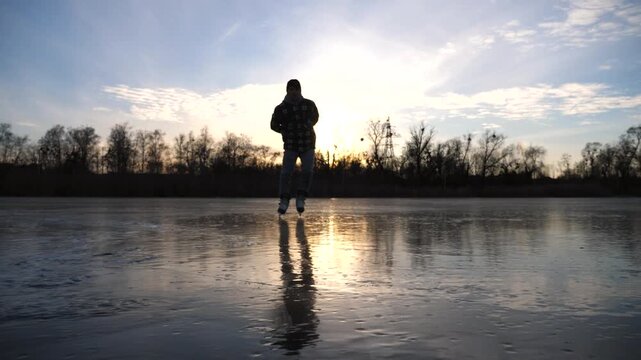 Guy skating on frozen river during sunset time. Young man shod in figure skates sliding on ice. Sun is reflecting on surface of icy lake. Male sportsman have active leisure at winter day. Slow motion