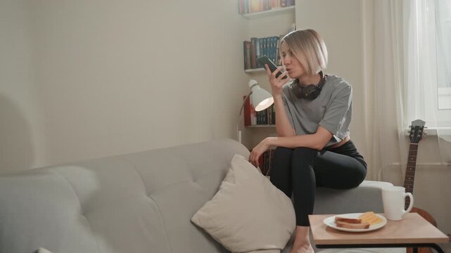 Wideshot caucasian woman sitting on couch with coffee and pastry, guitar nearby and headphones around neck, composing melody and sending soft serenade to lover, daylight through window creates cozy