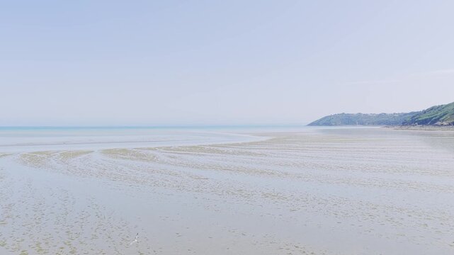 Wide shot of Binic beach on the Brittany coast of France during low tide, with wet sand patterns, shallow water, and a distant green headland under a clear blue sky.