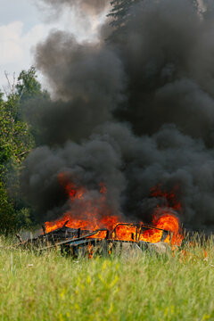 Vertical shot of a burning car wreck in a green grassy field with bright orange flames and huge black smoke column rising above trees on a summer day