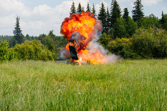 Huge fireball explosion of a car in a green grassy field with smoke and flying debris against a background of forest trees and cloudy sky on a summer day