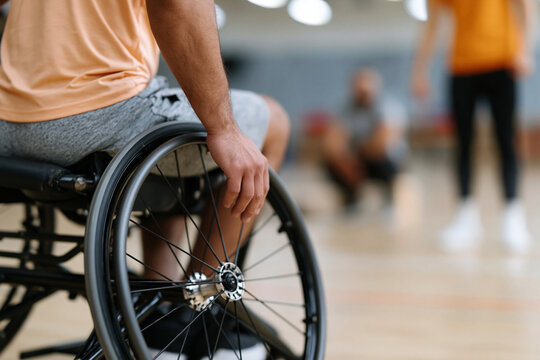 Man in wheelchair sits poised, focused. His hand rests lightly on the wheel's rim. Background hints at a gymnasium's bright space. Others move blurred, suggesting activity