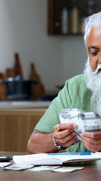 Indian mid age man counting Indian money notes at home, stylish bearded male managing monthly finance using calculator and diary while handling five hundred rupee currency notes at table