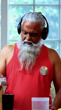 Indian mid age fit man with beard making protein shake post workout at home, stylish healthy male wearing headphones and sando vest preparing nutrition supplement in modern kitchen interior