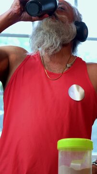 Indian mid age fit man with beard making protein shake post workout at home, stylish healthy male wearing headphones and sando vest preparing nutrition supplement in modern kitchen interior