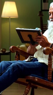 Indian mid age bearded man holds photo frame while sitting on rocking chair at home, stylish male with wet eyes recalling memories of loved one in modern interior emotional lifestyle moment