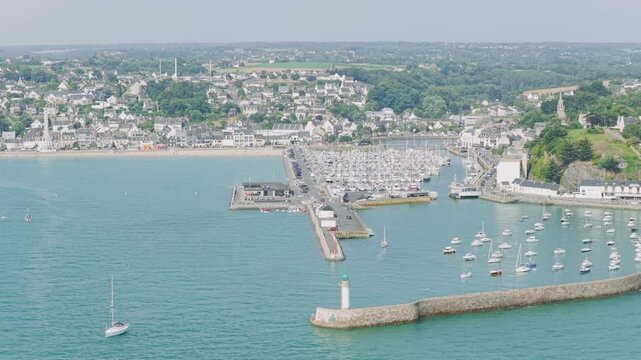 A high-angle aerial shot showcasing the marina of Binic with numerous docked sailboats and yachts, the protective harbor wall with a green and white lighthouse in Brittany, France.