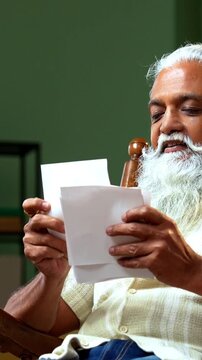 Indian mid age bearded man holds photographs prints while sitting on rocking chair at home, stylish male with wet eyes recalling precious memories of loved one in modern interior lifestyle
