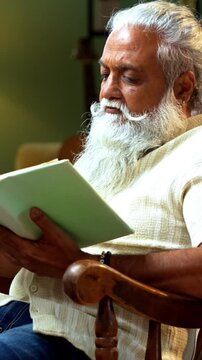 Indian mid age bearded man reading book novel on relaxing chair at home, stylish mature male enjoying peaceful leisure time with printed copy in modern interior lifestyle setting