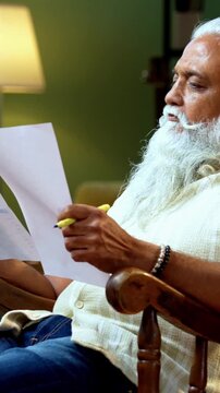 Indian mid age bearded man reading and showing financial document to camera sitting on relaxing chair at home, stylish mature male evaluating profit and loss report with positive expression