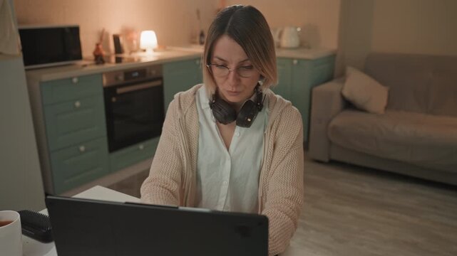 Evening scene of woman crafting melodies on laptop. Female musician focused on producing music during evening hours. Woman in cozy kitchen scene working on music with headphones and warm lighting