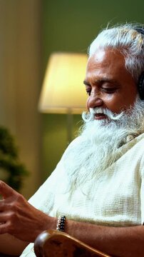 Indian mid age bearded man watching movie using smartphone sitting on relaxing chair at home, stylish mature male enjoying comedy short videos or reels with headphones in modern interior