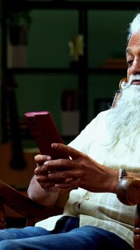 Indian mid age bearded man watching movie using smartphone sitting on relaxing chair at home, stylish mature male enjoying comedy short videos or reels with headphones in modern interior
