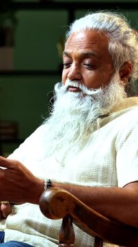 Indian mid age bearded man attending video call using smartphone sitting on relaxing chair at home, stylish mature male speaking during online video call in modern interior setting