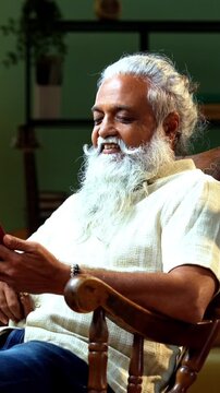 Indian mid age bearded man attending video call using smartphone sitting on relaxing chair at home, stylish mature male speaking during online video call in modern interior setting