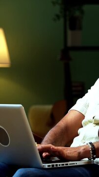 Indian mid age bearded man working on laptop sitting on relaxing chair at home, stylish mature male typing on computer in modern interior setting with wireless digital technology lifestyle