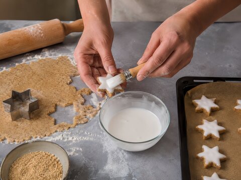 Zimtsterne werden von Hand mit wei&szlig;er Glasur bestrichen: H&auml;nde verzieren sternf&ouml;rmige Pl&auml;tzchen vor dem Backen &ndash; traditioneller deutscher Weihnachtsklassiker f&uuml;r Advent und Festtage.