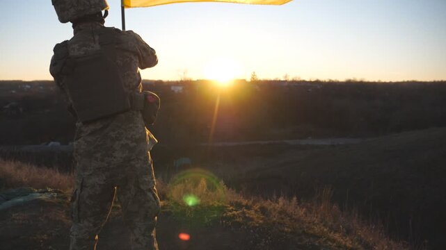 Ukrainian army man stands at the peak of hill waving blue-yellow banner against sunset. Male soldier in camouflage uniform lifting flag of Ukraine as a symbol of victory against russian aggression