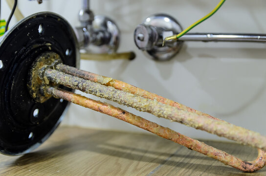 Heavily scaled water heater immersion heating element removed, showing thick limescale buildup from hard water, next to chrome plumbing valves
