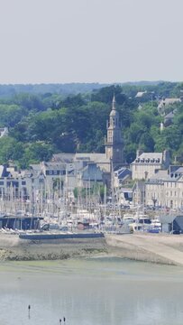 A wide, sunlit view of Binic, a coastal town in Brittany, France, showing the marina with moored sailboats, historic stone buildings, and church tower.