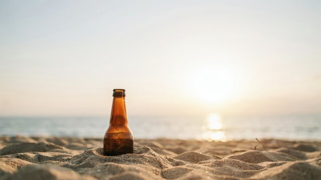 Brown glass beer bottle standing in sand on a beach at sunset