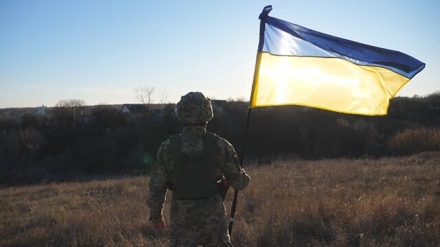Rear view of male soldier in military uniform holds a waving national flag. Ukrainian army man stands with lifting blue-yellow banner at field. End of war in Europe. Invasion of territory Ukraine