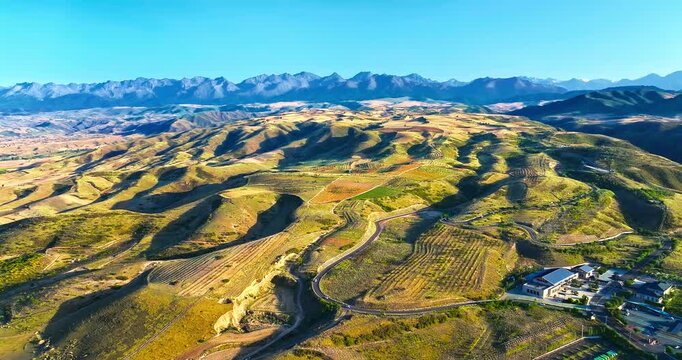 Rolling cultivated hills, winding roads, and rural buildings stretch toward a jagged mountain range, highlighting agricultural landscape and regional tourism in Xinjiang, China.