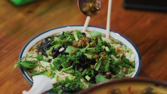 Closeup noodle bowl being finished with sauce and herbs, chopstick and spoon in motion over wooden table, scallion and sesame topping, steaming broth and savory umami aroma, comforting kitchen plating