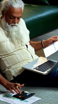 Indian mid age bearded man using laptop, calculator and reports for accounting sitting on carpet in modern home, stylish mature male managing finance, budget and financial statements indoors