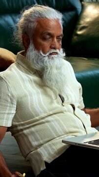 Indian mid age bearded man using laptop, calculator and reports for accounting sitting on carpet in modern home, stylish mature male managing finance, budget and financial statements indoors
