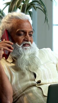 Indian mid age bearded man working online using laptop, smartphone call and reports sitting on sofa in modern home, stylish mature male multitasking business discussion and paperwork indoors