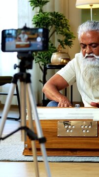 Indian mid age bearded man recording harmonium playing session on smartphone for online class at home, stylish mature music tutor creating classical lesson video with tripod setup in modern studio