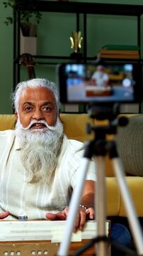 Indian mid age bearded man recording vocals music session on smartphone for online class at home, stylish mature tutor teaching Indian classical singing with harmonium in modern studio