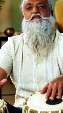 Indian mid age stylish bearded man taking online Tabla musical instrument playing class at home, experienced classical percussion tutor streaming lesson on smartphone in modern studio