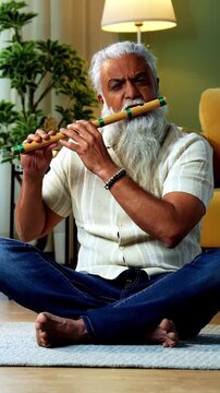 Indian mid age stylish bearded man teaching playing wooden flute musical instrument at home using smartphone, experienced classical bansuri tutor conducting online class in modern studio