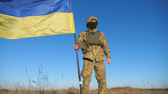 Ukrainian army man in military uniform lifting national banner. Young male soldier holds a waving flag of Ukraine against blue sky. Victory against russian aggression. Invasion resistance. Low view
