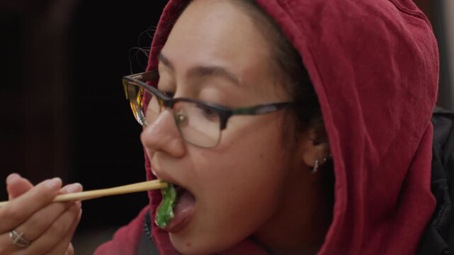 Hooded asian woman nighttime pause with chopsticks and thoughtful gaze closeup portrait under soft neon glow, glasses catching bokeh, quiet street stall atmosphere, reflective mood after evening walk