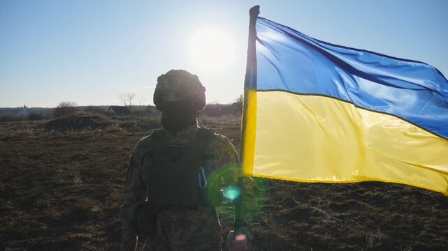 Soldier of ukrainian army stands with lifting blue-yellow banner at field. Male military in camouflage uniform holds a waving flag of Ukraine. Victory against russian aggression. Invasion resistance