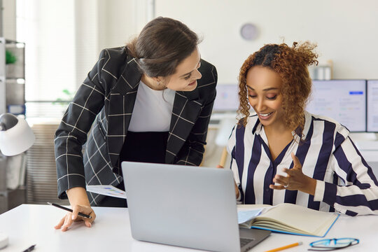 Businesswomen coworkers in office discuss planning over laptop. They share ideas, review notes and data, align on project goals, showing collaboration and teamwork. Clear team plan concept message.