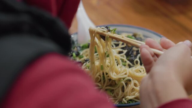 Close up hands twirling noodles bowl, chopsticks lifting strands over ceramic bowl with scallion and mushroom garnish, wooden table, red jacket sleeve, candid cozy meal moment with texture and steam