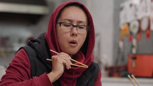 Late night diner woman eating noodles, cozy urban eatery, wearing red hoodie and scarf, using chopsticks, thoughtful expression, warm ambient lighting, menu board in background, candid tasting moment,