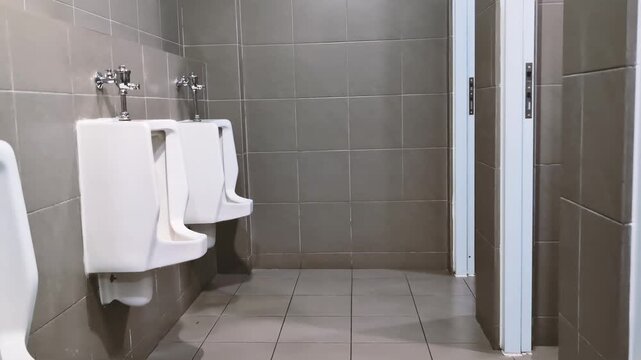 Clean modern empty men public restroom toilet corner interior featuring white ceramic urinal on contemporary grey tiled wall alongside closed stall door providing sanitary hygiene facility space
