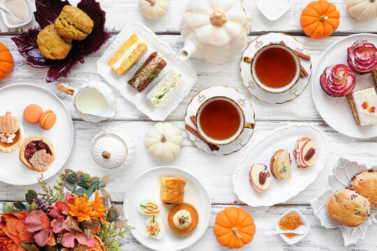 Autumn theme afternoon tea table scene over a white wood background. Pumpkin and apple theme savory finger foods, sandwiches, scones, and desserts. Top view.